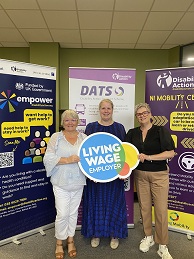 Three people standing facing the camera. They are standing infront of three popup stands holding a Living Wage Employer sign.