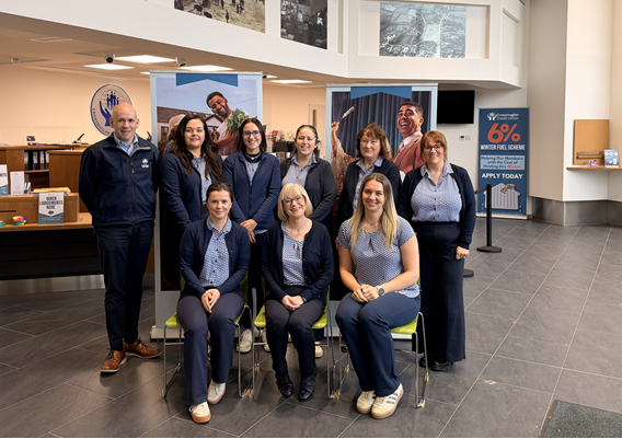 Nine people from Crossmaglen Credit Union are in a large room. Some are sitting and others are standing. They are looking at the camera and smiling.
