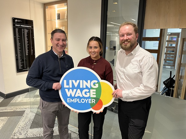 Three people are standing in a modern building. They are holding a "Living Wage Employer" sign and smiling at the camera.