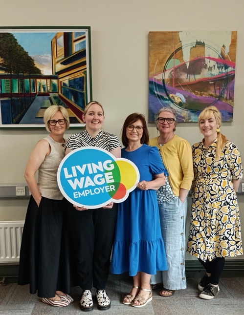 Five women are standing in a row. They are dressed casually and smiling at the camera in front of a wall with two brightly coloured pictures.