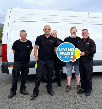 Four people dressed in black standing in front of a white van and holding the Living Wage Employer sign.