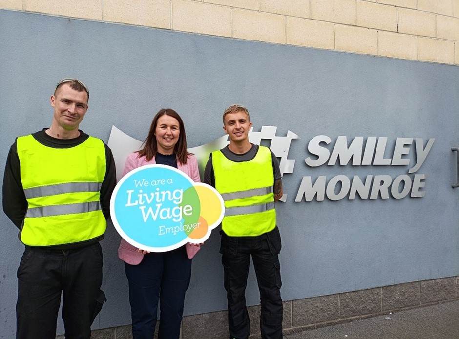 Three people standing in front of the Smiley Monroe logo holding a sign that states "We are a living wage employer".