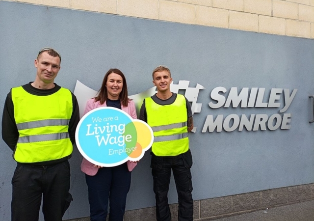 Three people standing in front of the Smiley Monroe logo holding a sign that states "We are a living wage employer".