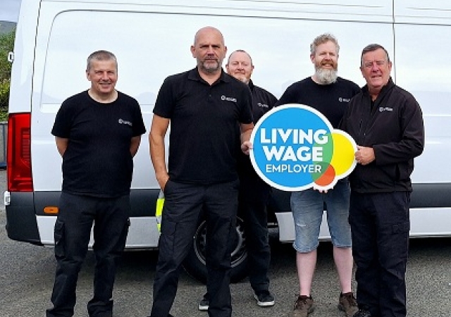Four people dressed in black standing in front of a white van and holding the Living Wage Employer sign.