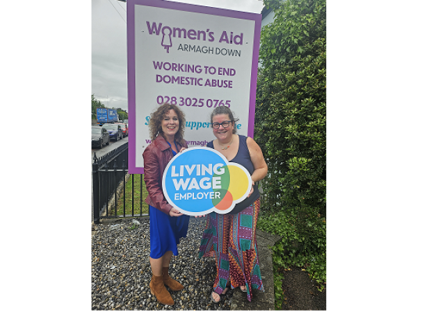 Two women standing in front of the Women's Aid pop-up standing holding a 'Living Wage Employer' sign.