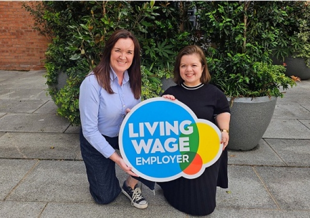 Two people kneeling down in front of green plants and holding a Living Wage Employer sign.